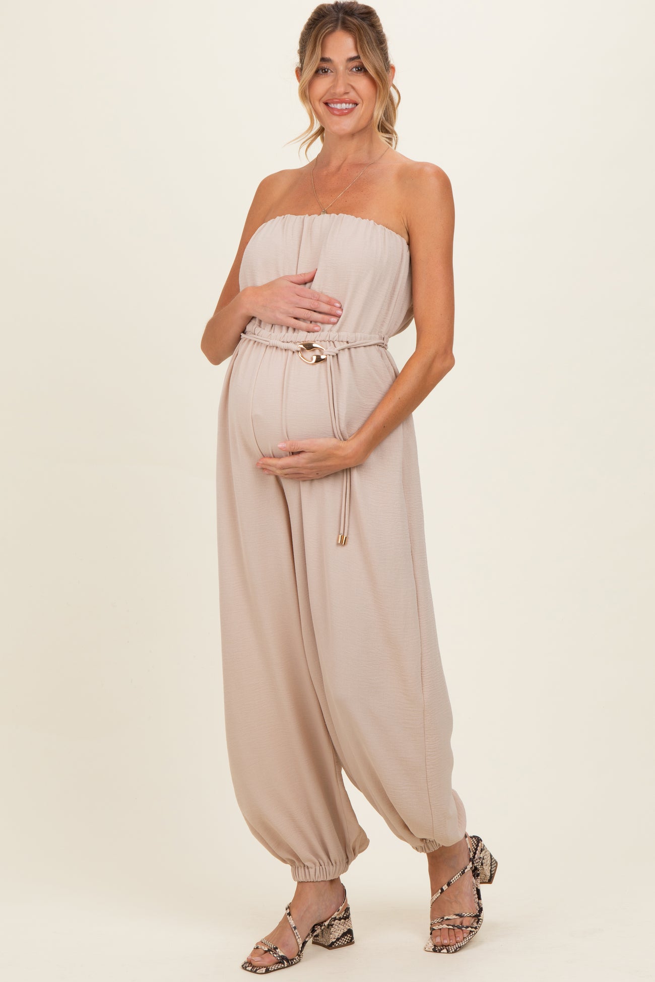 woman wearing a beige strapless maternity jumpsuit with a cinched waist and holding her baby bump