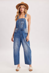 woman wearing a light blue denim overall with wide legs and front pockets, styled with a white tank top and a straw hat