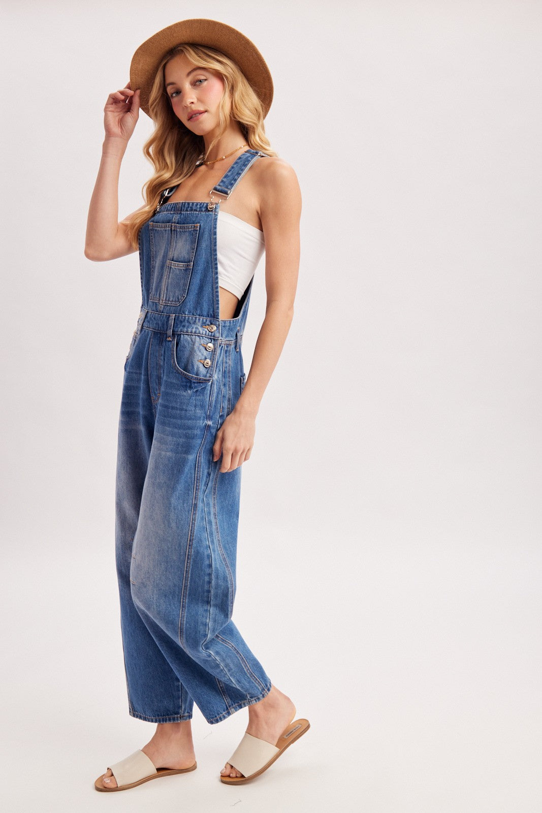 woman wearing a light wash denim overall with wide legs, paired with a white tank top and straw hat