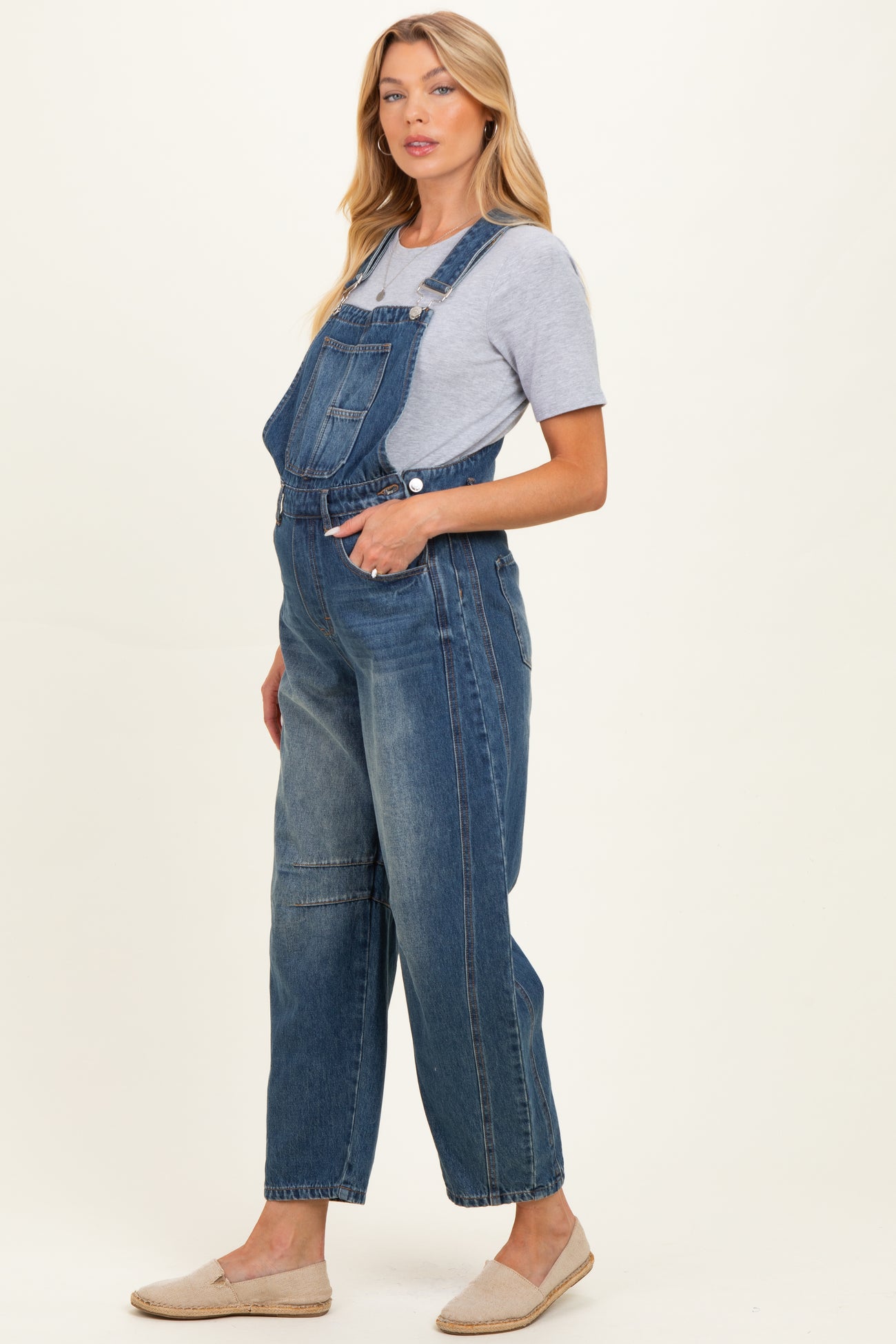 woman wearing a light blue maternity denim overalls with a grey t-shirt underneath and casual slip-on shoes