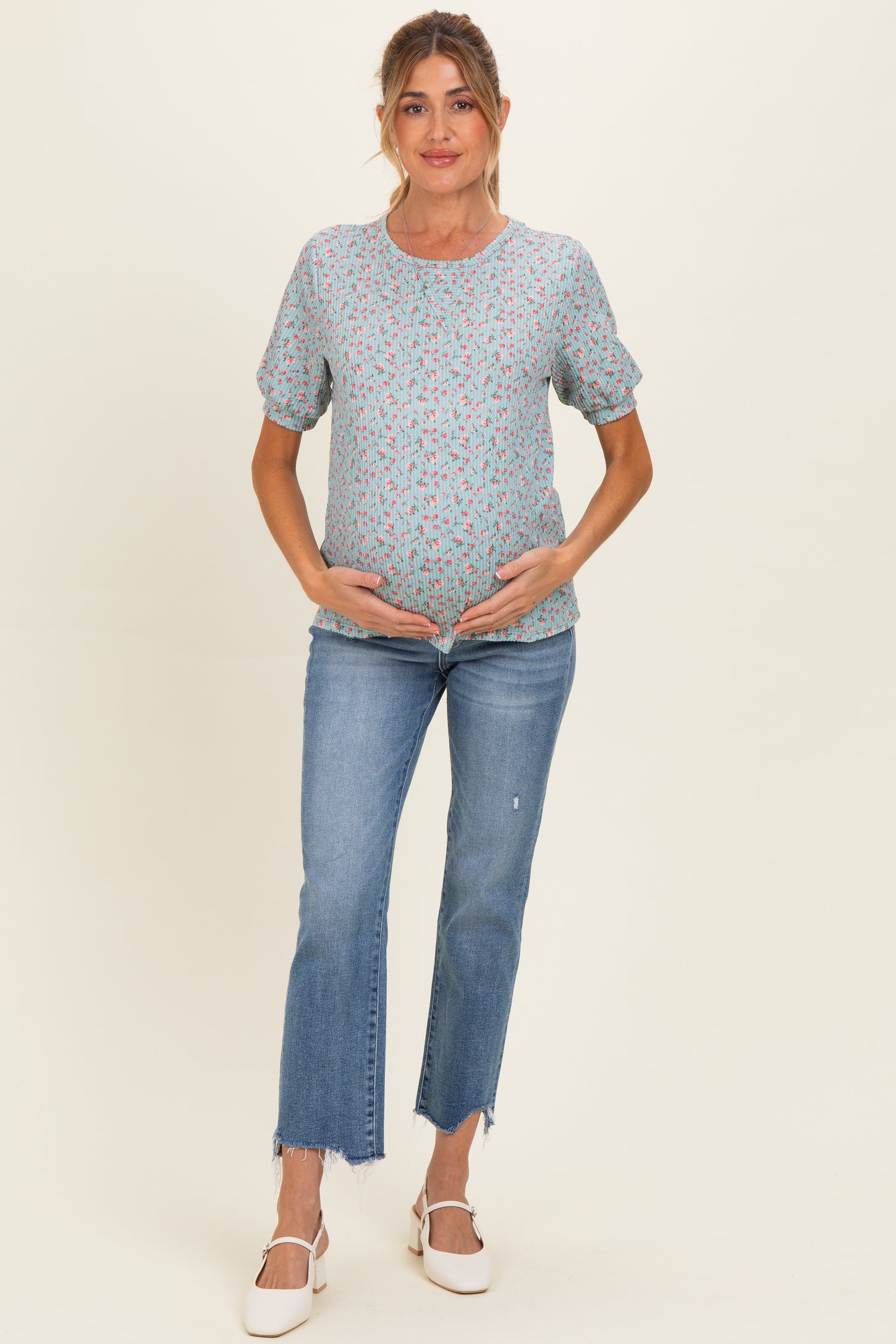 woman wearing a light blue maternity top with floral print and holding her baby bump, paired with cropped jeans