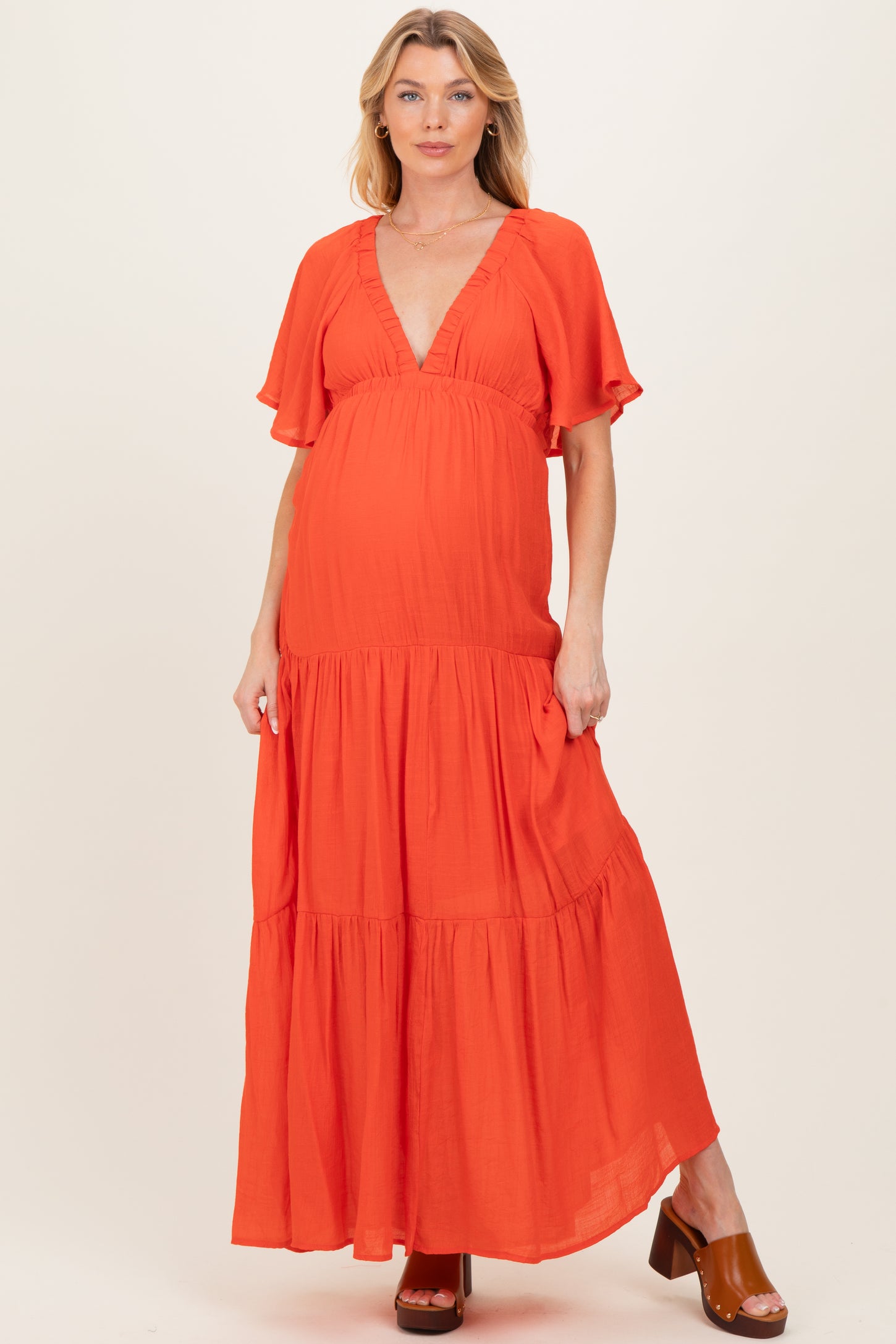 woman wearing a vibrant orange maternity maxi dress with a deep v-neckline and flutter sleeves, showcasing her baby bump