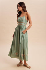 woman wearing a green maxi dress with a gathered waist and palm tree embroidery, standing in profile