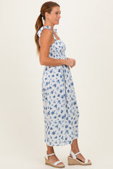 woman wearing a white floral midi dress with blue patterns and tied shoulder straps, standing sideways