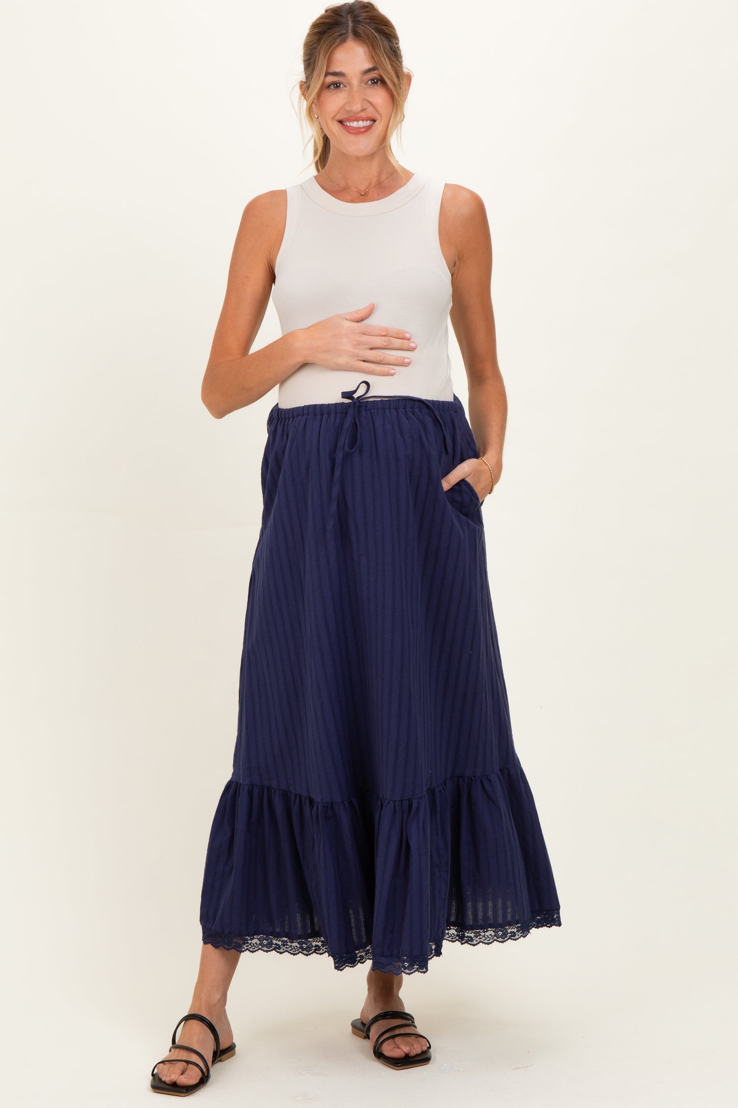 woman wearing a cream sleeveless maternity top and a dark blue tiered maxi skirt, holding her baby bump