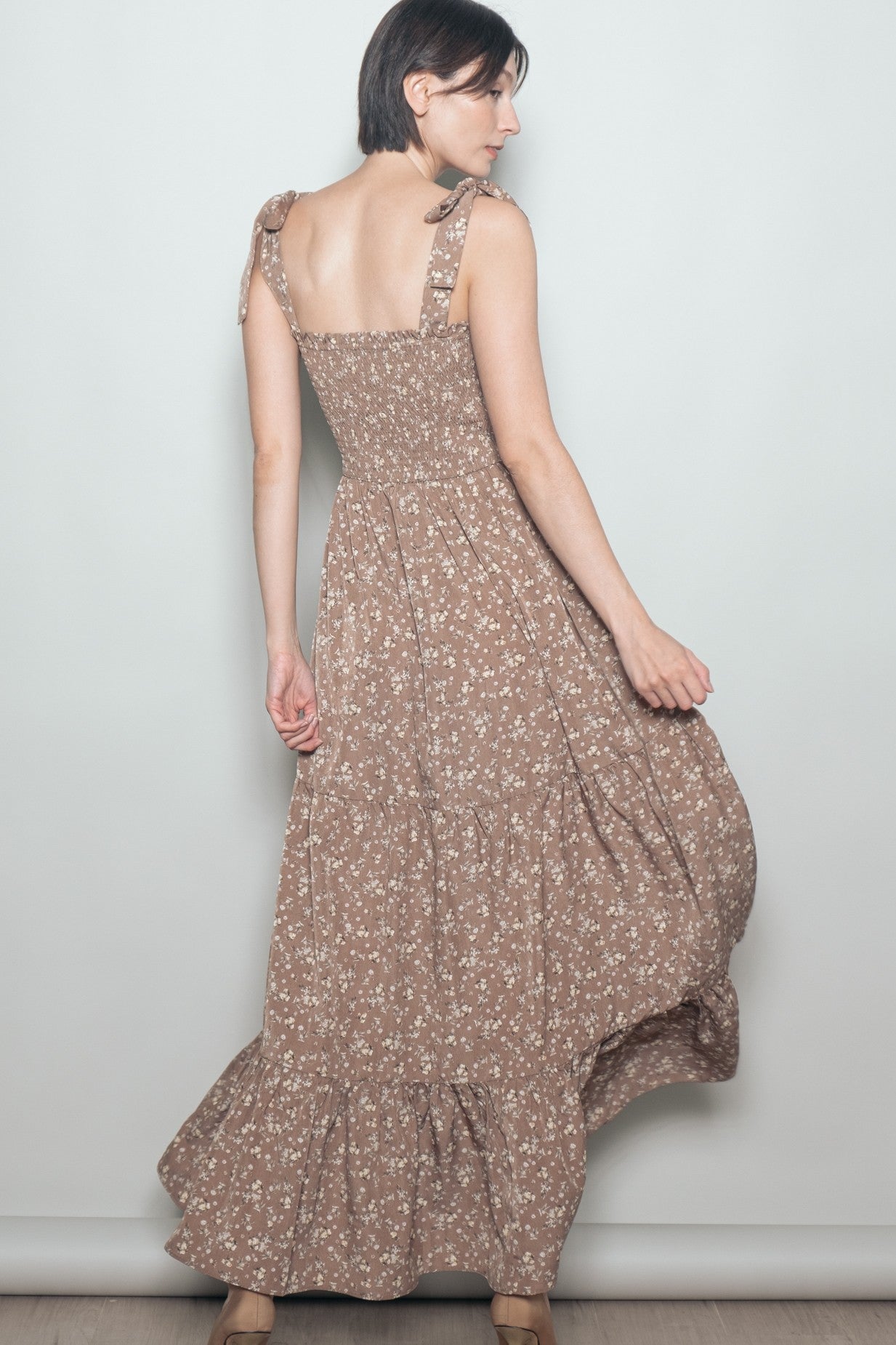 woman wearing a brown floral maxi dress with tie straps and a tiered skirt, posing with her back to the camera