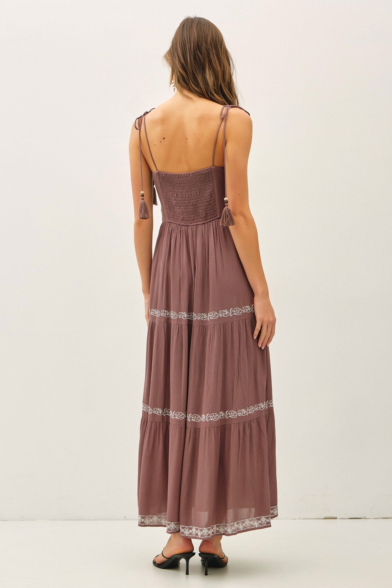 woman wearing a mauve maxi dress with a smocked bodice and tassel straps, showing the back view