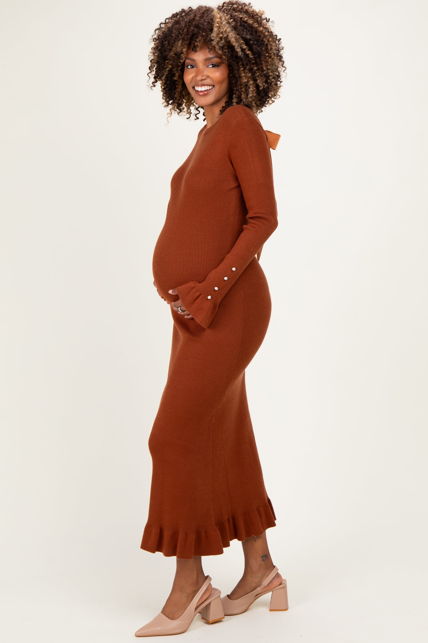 woman wearing a rust brown long sleeve ribbed maternity dress with a ruffle hem and a bow detail on the back, holding her baby bump