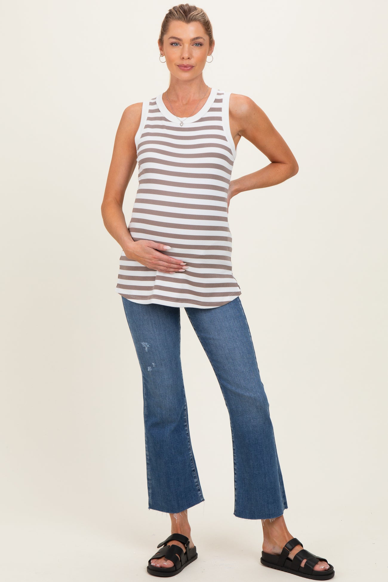 woman wearing a gray and white striped maternity tank top and distressed blue flared jeans, gently holding her baby bump