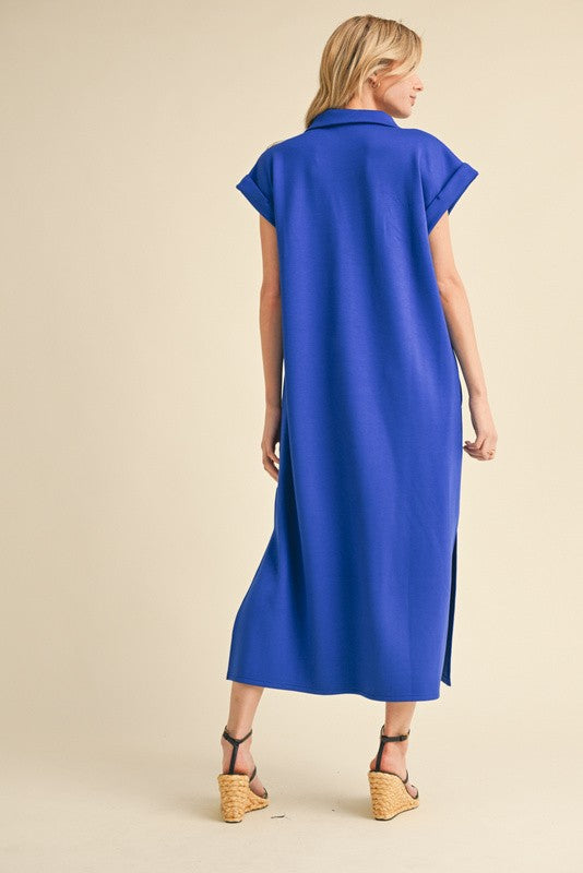 woman wearing a blue short-sleeve maxi dress with a high slit and back collar detail, standing against a beige background