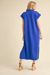 woman wearing a blue short-sleeve maxi dress with a high slit and back collar detail, standing against a beige background