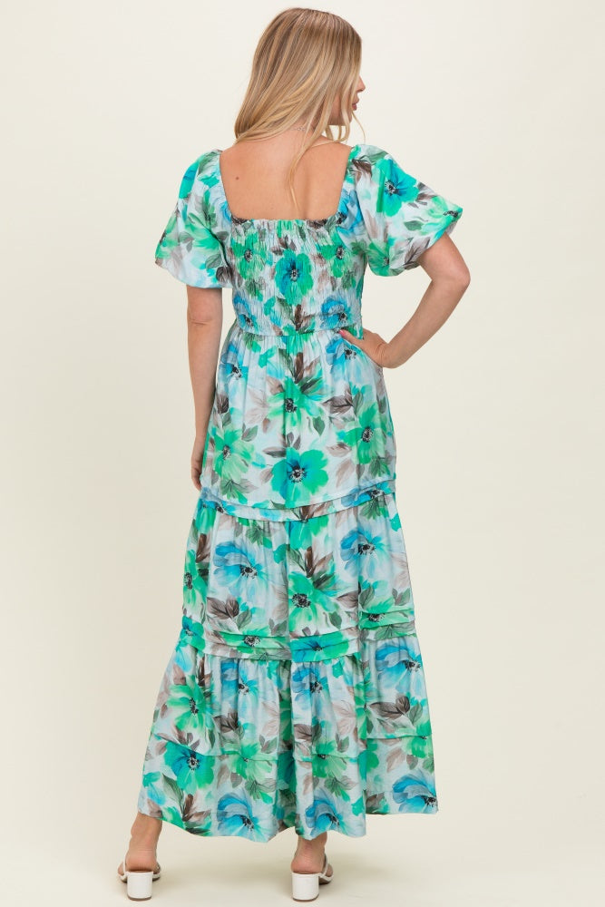 woman wearing a flowy maxi dress with a floral print in shades of blue and green, featuring a smocked bodice and tiered skirt