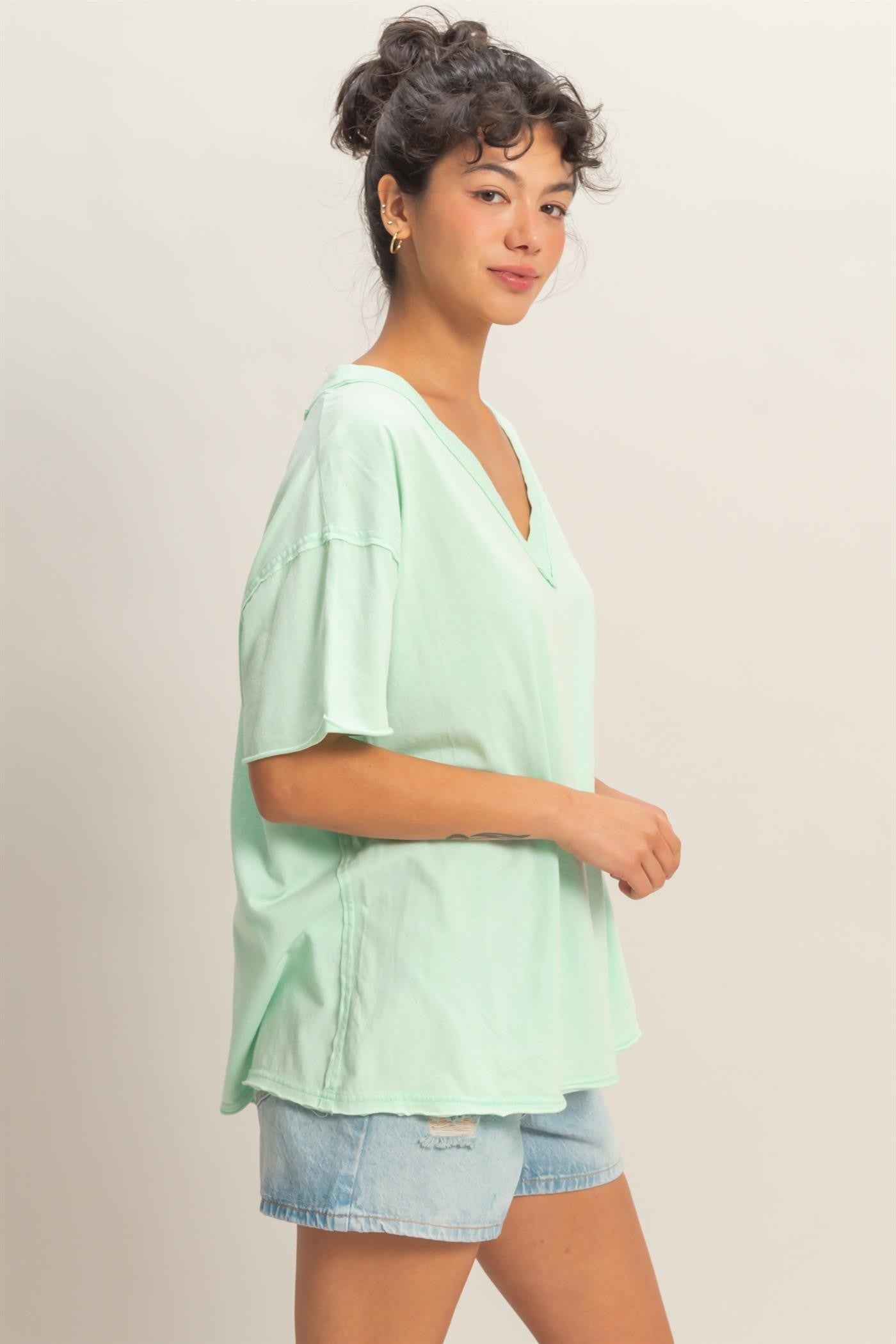 woman wearing a mint green oversized v-neck t-shirt and denim shorts, standing sideways with a slight smile