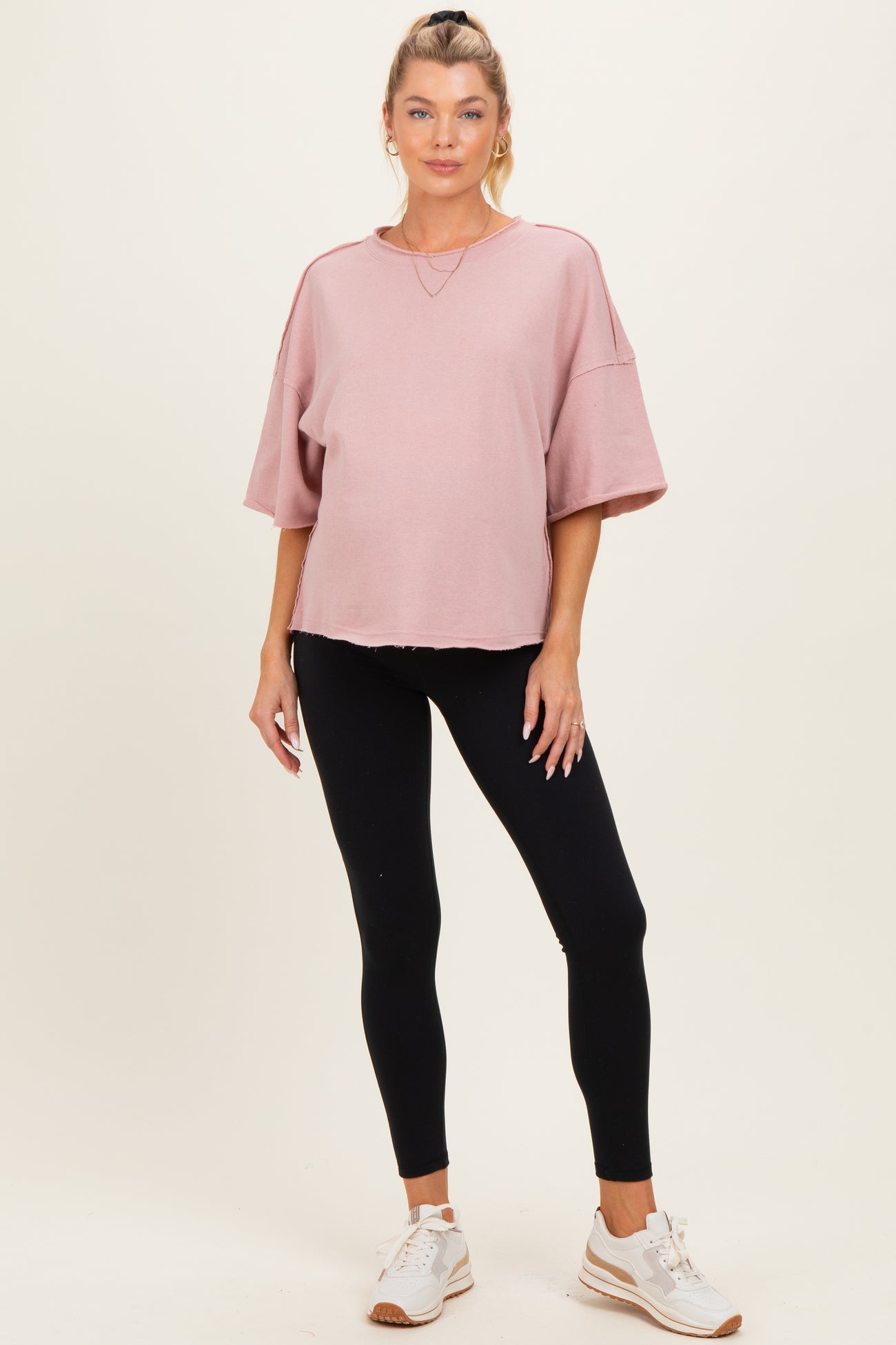woman wearing a pink oversized maternity top and black leggings, standing confidently on a light background