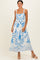 woman wearing a white maxi dress with blue floral print and scalloped hem, standing against a neutral background
