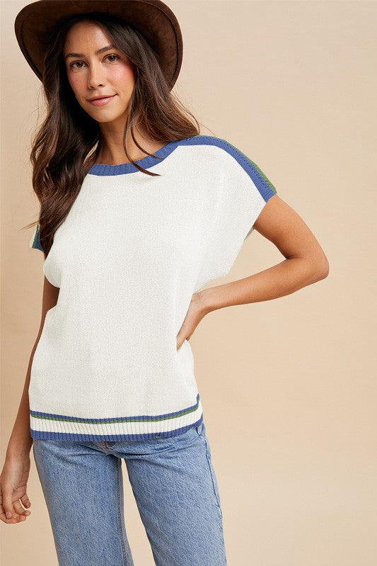 woman wearing a white short sleeve sweater with blue and green accents, paired with light blue jeans and a brown hat