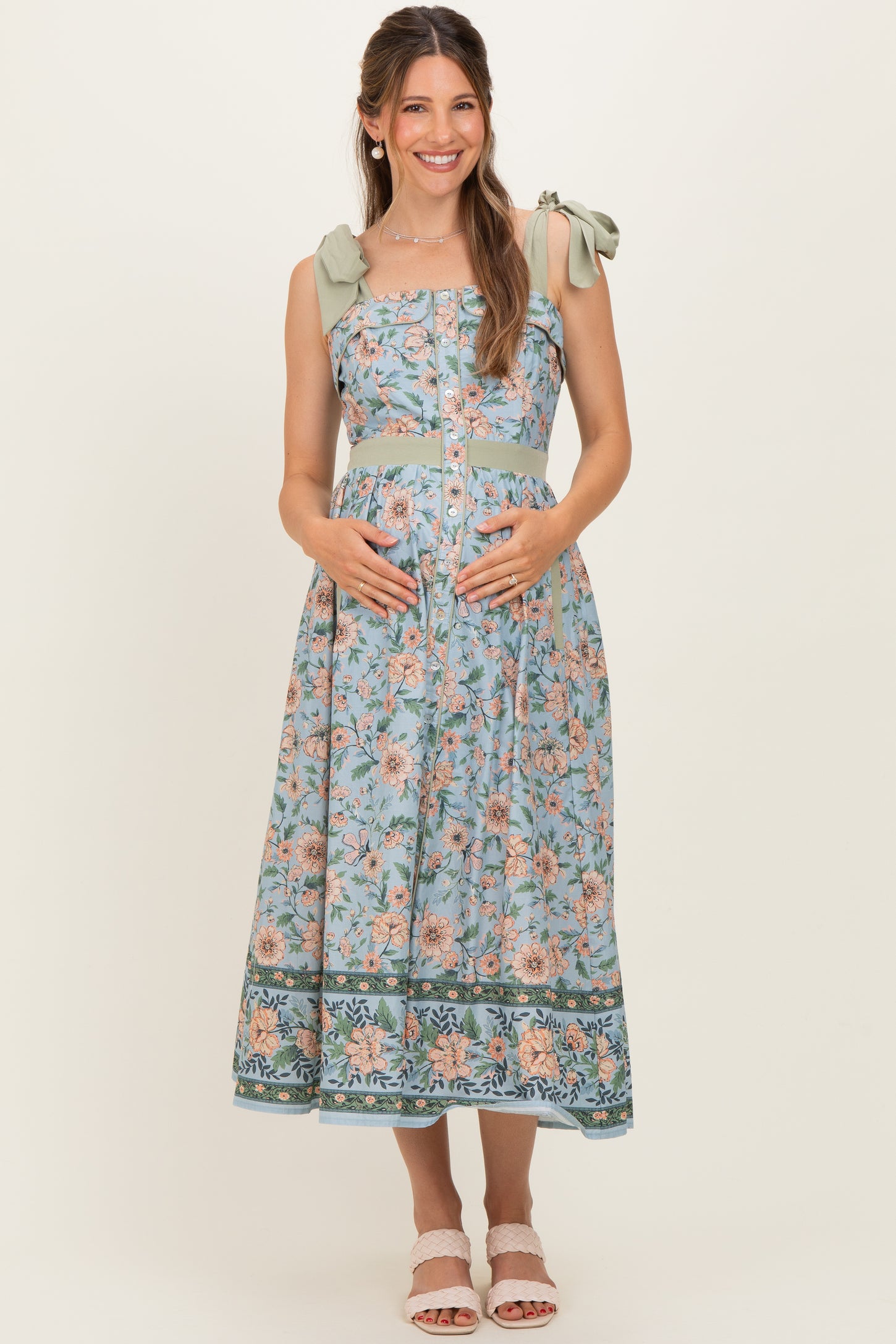 woman wearing a floral print maternity dress with bow straps and a baby bump visible, standing and smiling