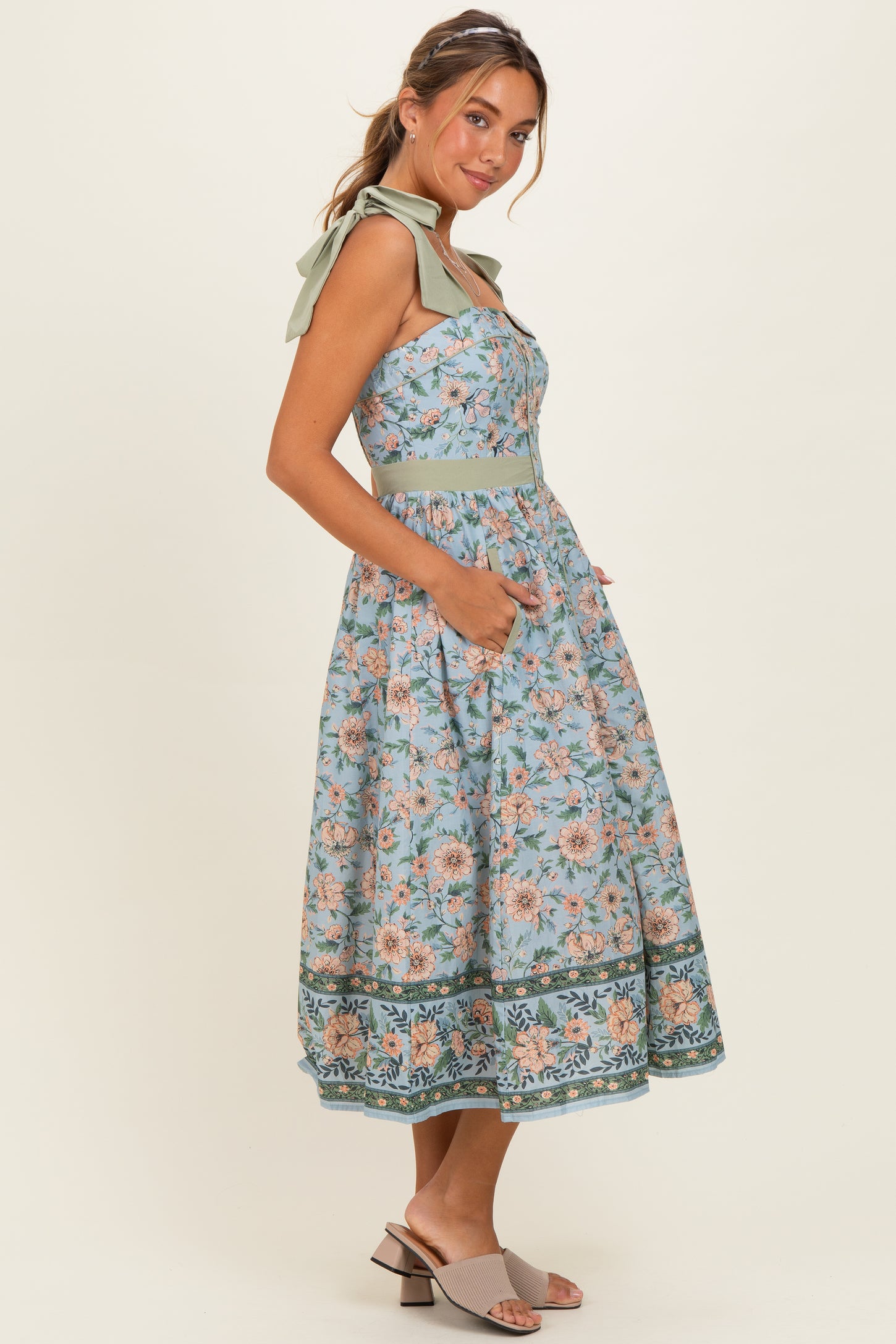 woman wearing a blue floral midi dress with bow straps and pockets, styled with gray sandals