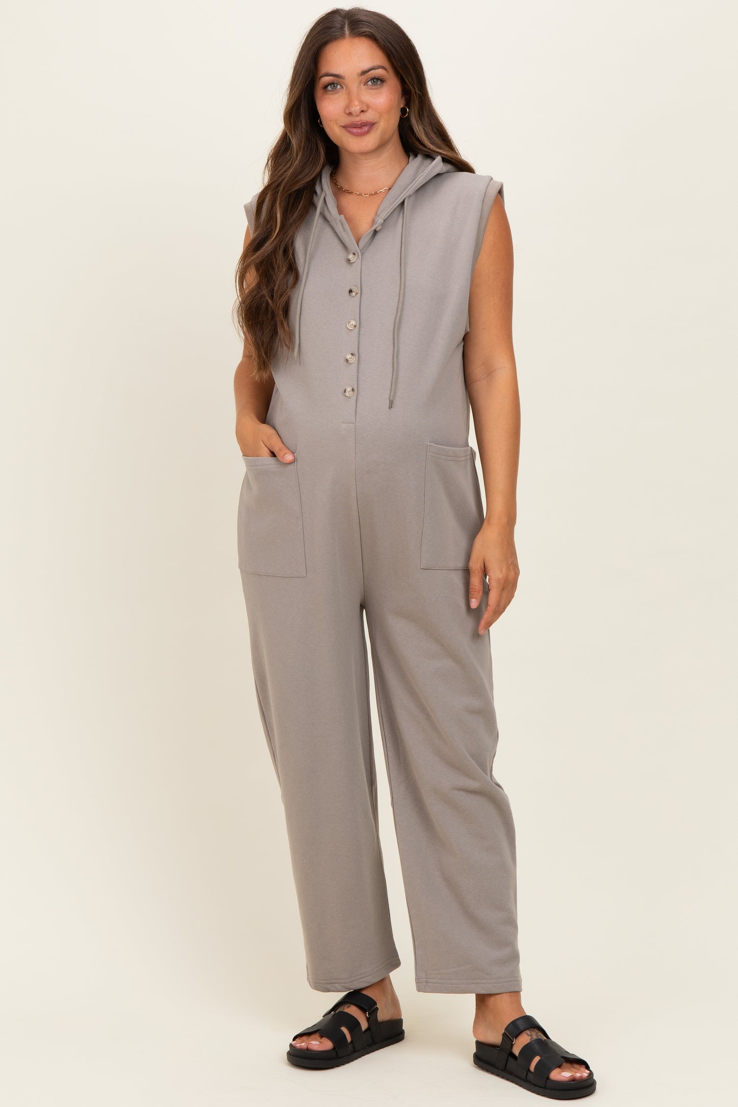 woman wearing a gray sleeveless maternity jumpsuit with hood and pockets, styled with sandals