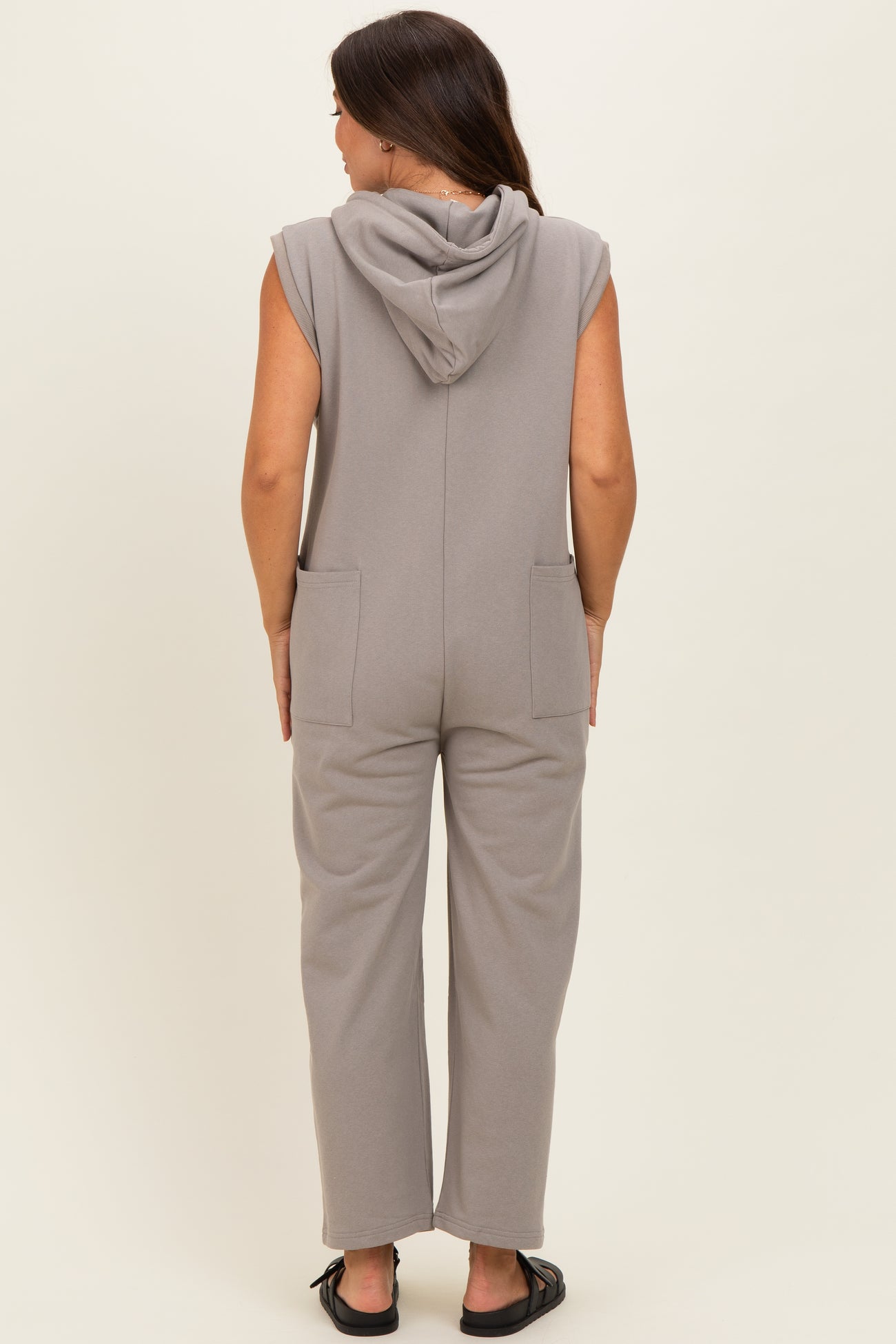 woman wearing a grey sleeveless maternity jumpsuit with a hood and pockets, shown from the back