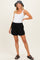 woman wearing a white maternity tank top and black maternity shorts, posing with a smile and a hat