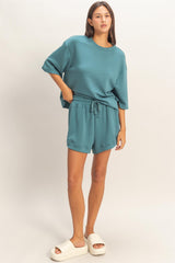woman wearing a teal oversized sweatshirt and matching shorts with a drawstring, paired with cream slide sandals
