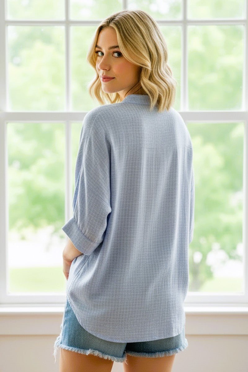 woman wearing a light blue oversized button-up shirt with rolled sleeves and denim shorts, standing by a window