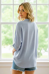 woman wearing a light blue oversized button-up shirt with rolled sleeves and denim shorts, standing by a window