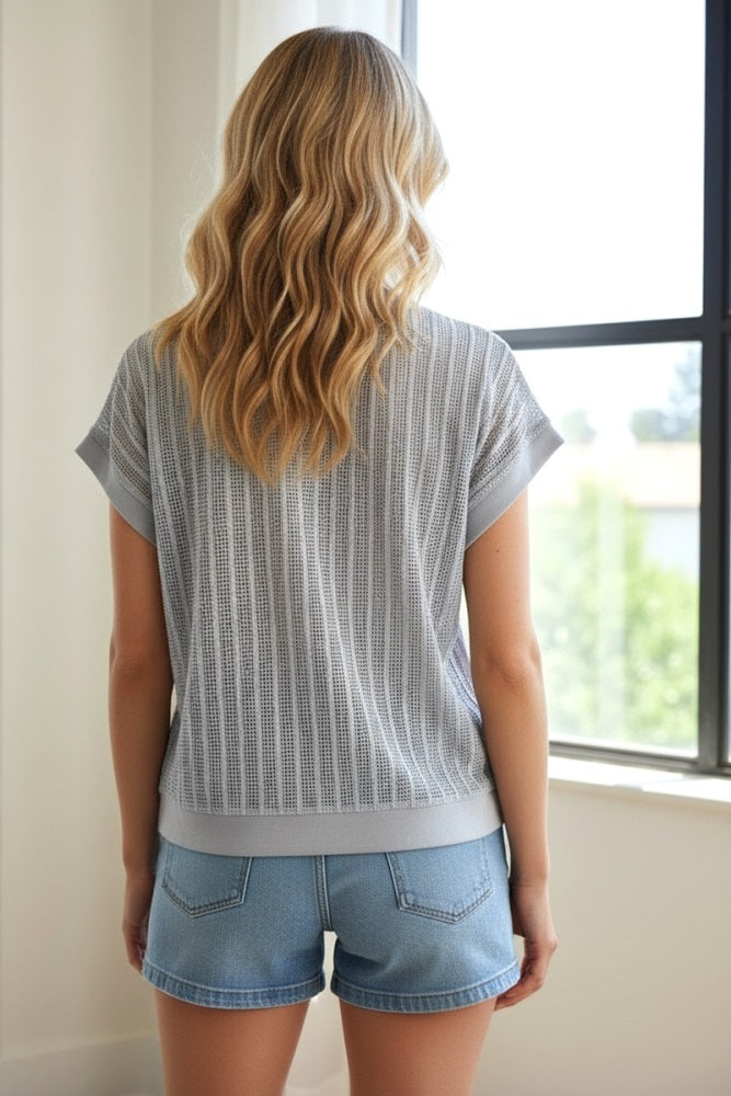 woman wearing a light gray patterned short sleeve top and denim shorts, seen from the back near a window