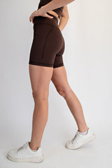 woman wearing brown high-waisted workout shorts and white sneakers standing in profile with a plain background
