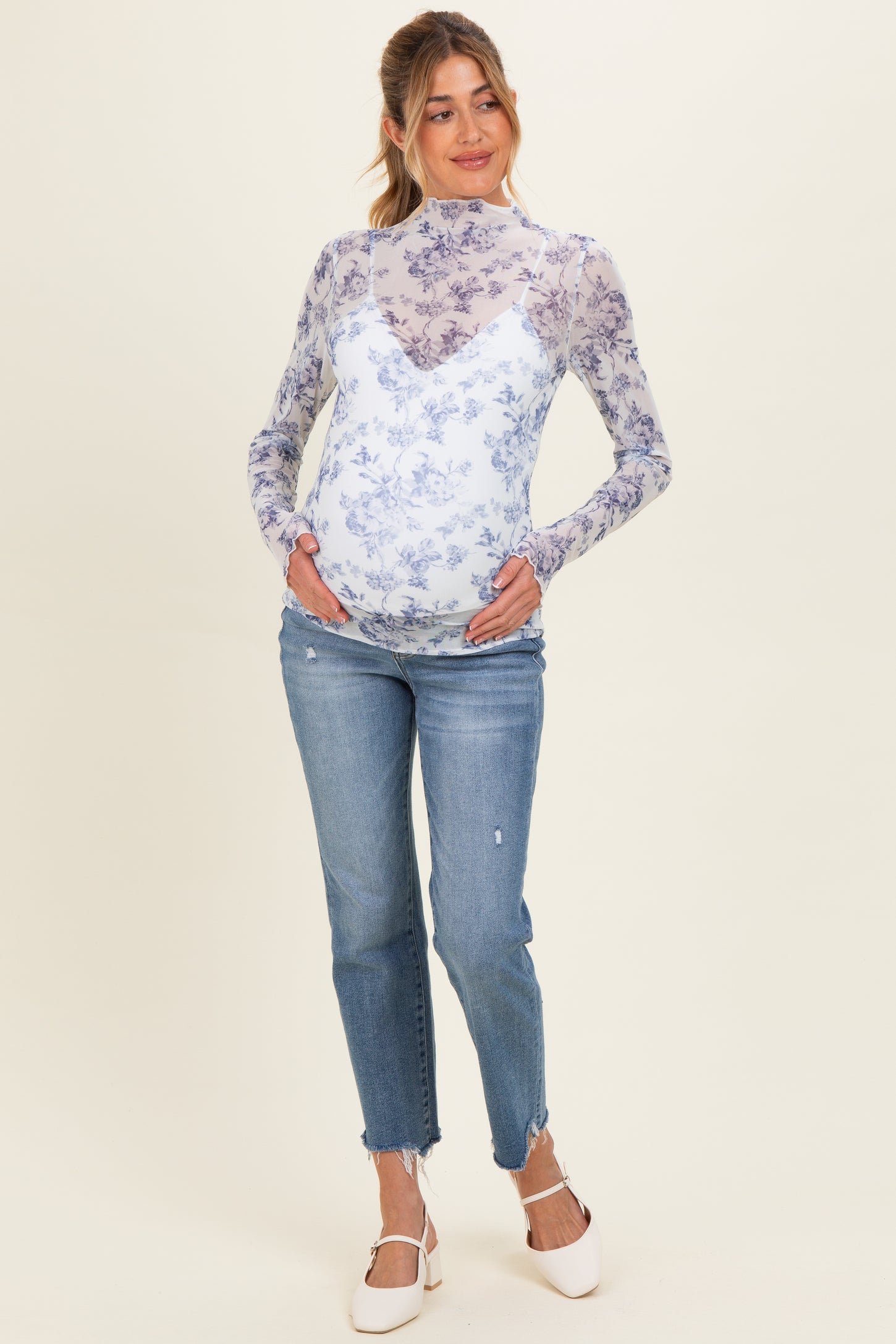 woman wearing a blue floral print maternity top with a sheer overlay and light blue jeans, cradling her baby bump
