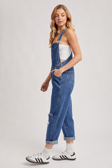 woman wearing a pair of distressed denim overalls with a white tank top and black and white sneakers