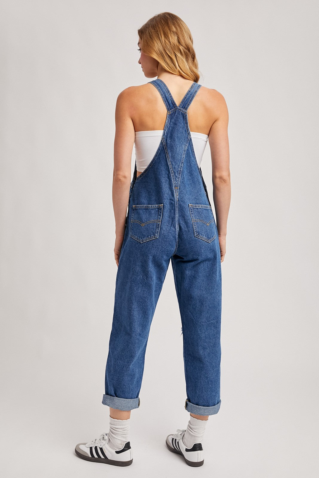 woman wearing a blue denim overall with rolled cuffs and a white tank top, viewed from the back