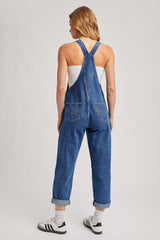 woman wearing a blue denim overall with rolled cuffs and a white tank top, viewed from the back