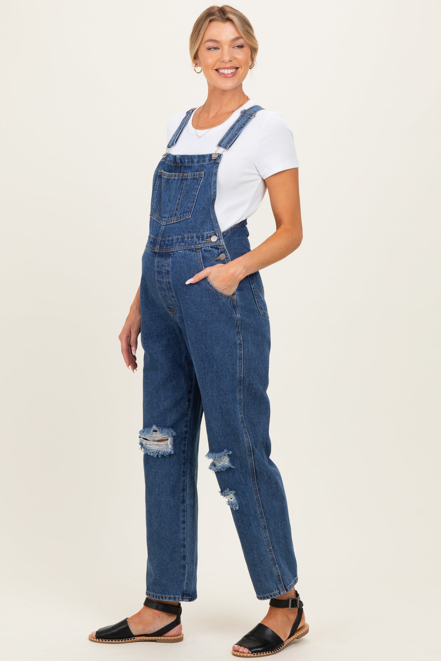 woman wearing maternity denim overalls with distressed details and a white t-shirt, standing sideways with a relaxed fit