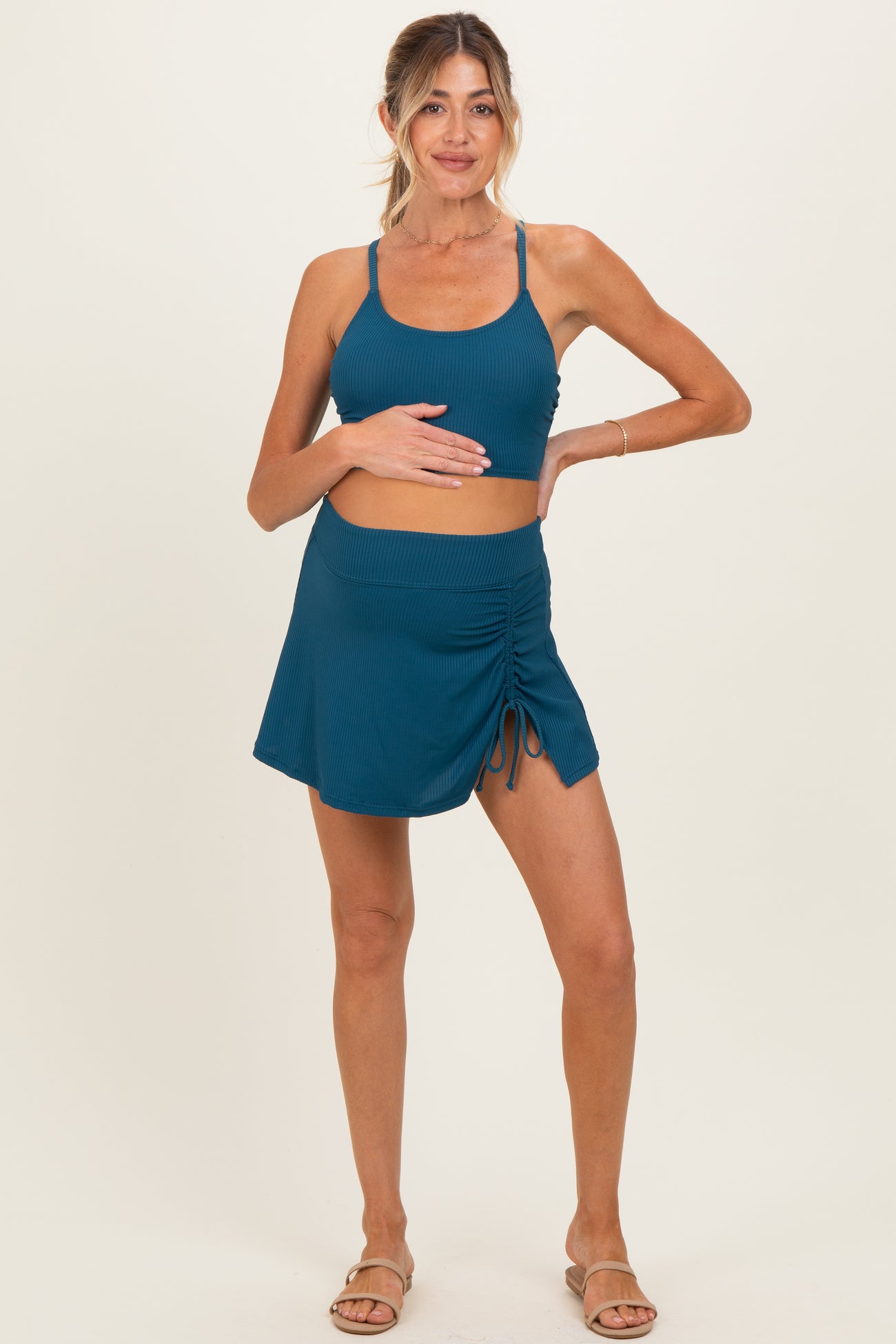 woman wearing a teal maternity crop top and a matching ruched skirt, gently holding her baby bump