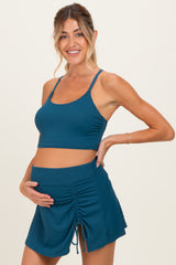 woman wearing a teal maternity crop top and matching skirt, holding her baby bump with a smile