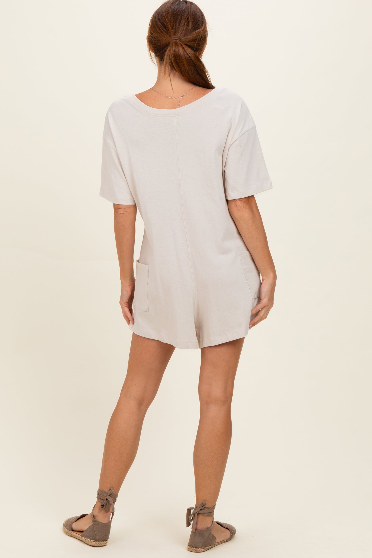 woman wearing a light beige short sleeve romper with a relaxed fit and side pockets, view from the back