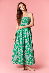 woman wearing a green maxi dress with a floral print and smocked bodice, posed against a pink background