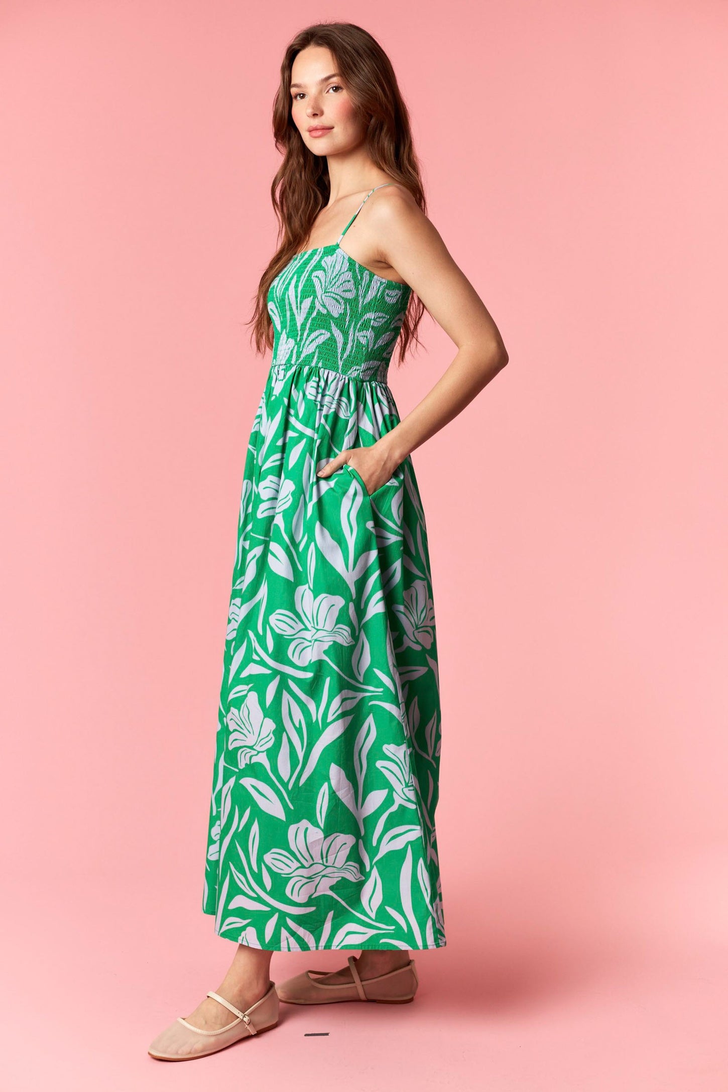 woman wearing a green floral maxi dress with a fitted bodice and pockets, standing against a pink background