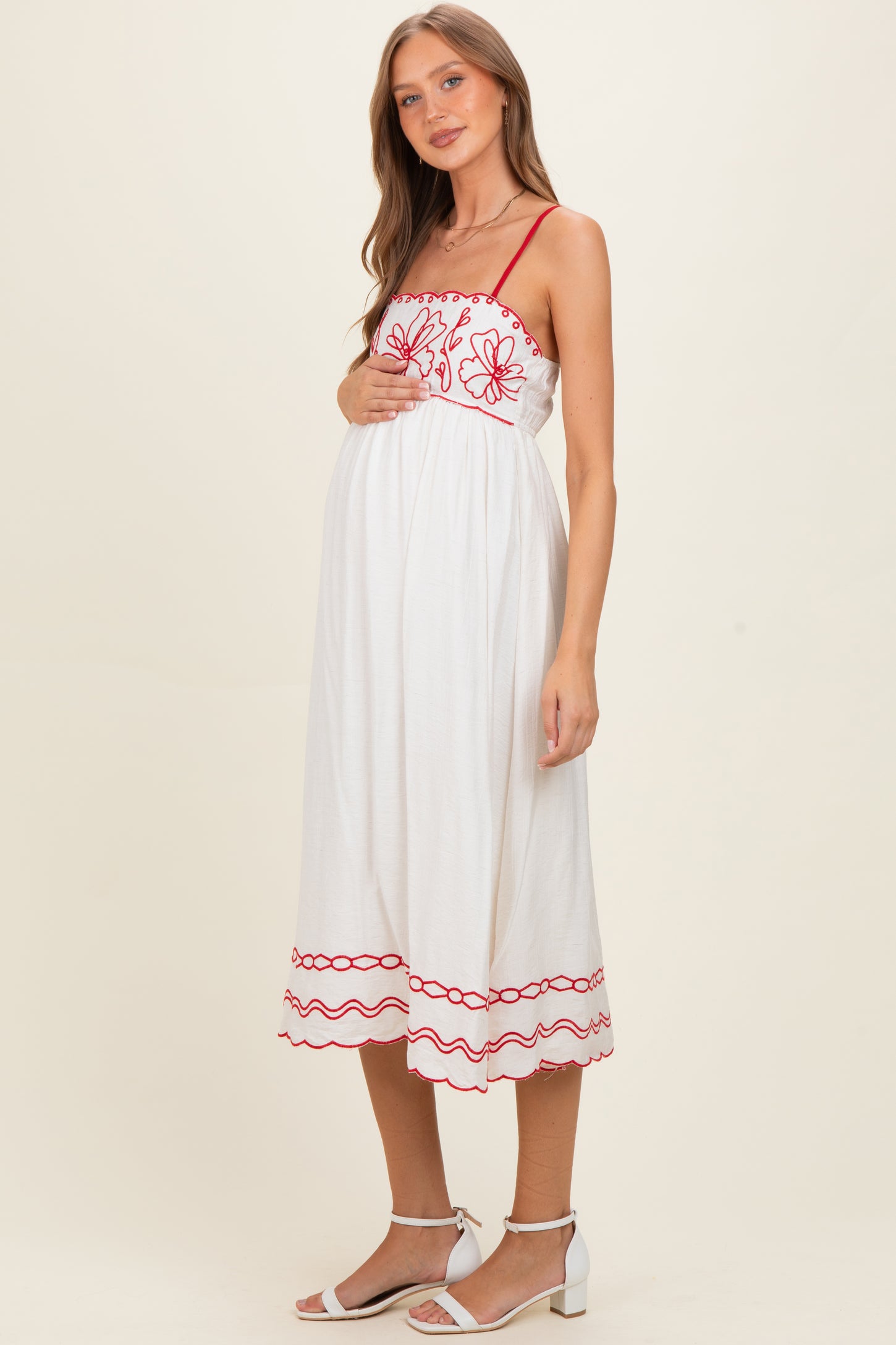 woman wearing a white maternity dress with red floral embroidery and spaghetti straps, highlighting her baby bump