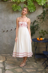 woman wearing a white midi dress with red embroidery details and spaghetti straps, standing outdoors among greenery