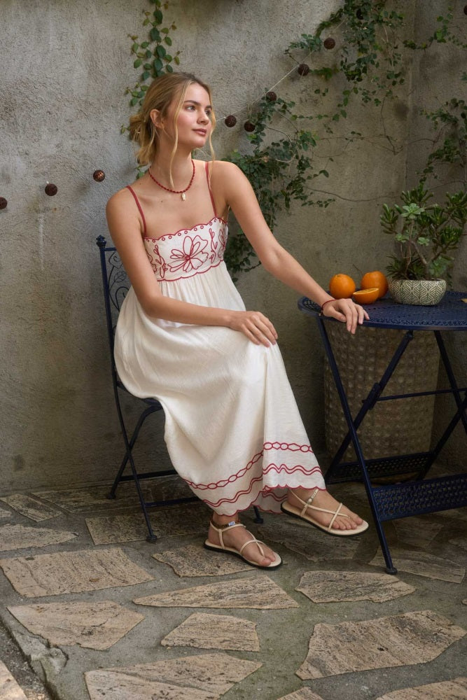 woman wearing a white maxi dress with floral embroidery sitting on a patio chair, sandals on her feet