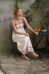 woman wearing a white maxi dress with floral embroidery sitting on a patio chair, sandals on her feet