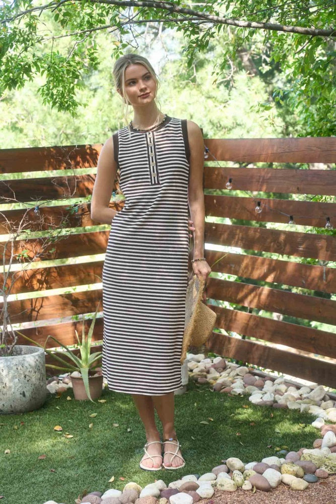 woman wearing a striped sleeveless maxi dress with a button detail, standing outdoors among greenery and rocks