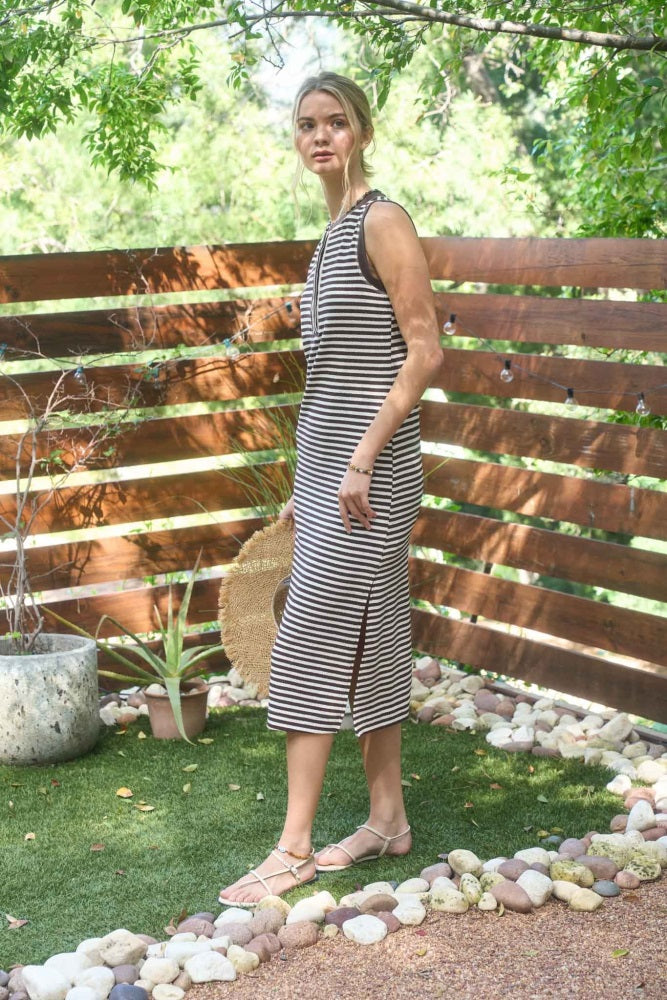 woman wearing a striped sleeveless midi dress with a side slit, standing outdoors with greenery in the background