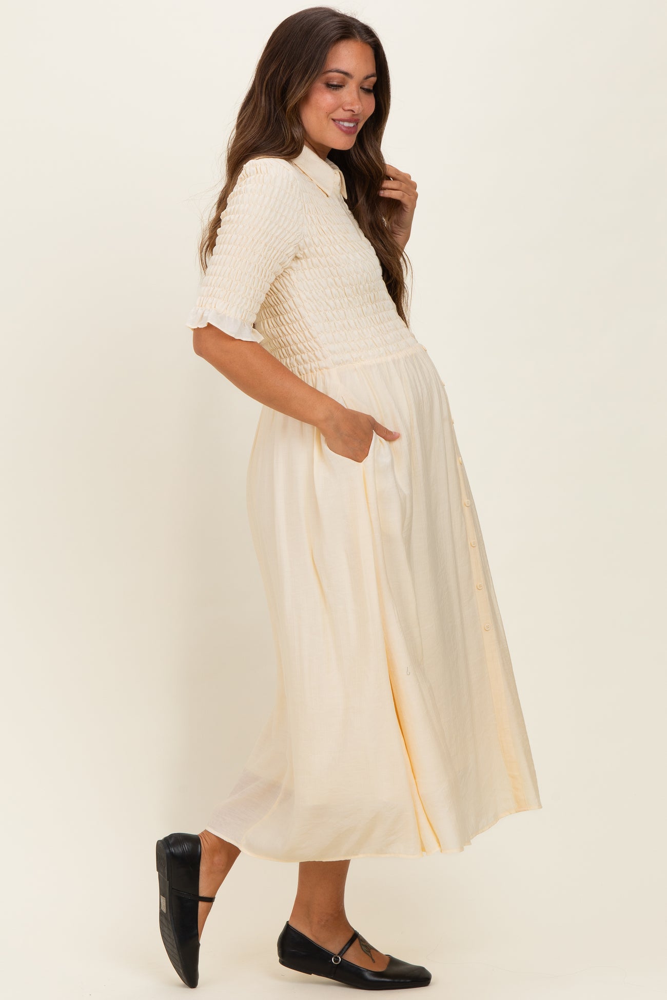 woman wearing a cream-colored maternity dress with a smocked bodice and side pockets, showcasing her baby bump