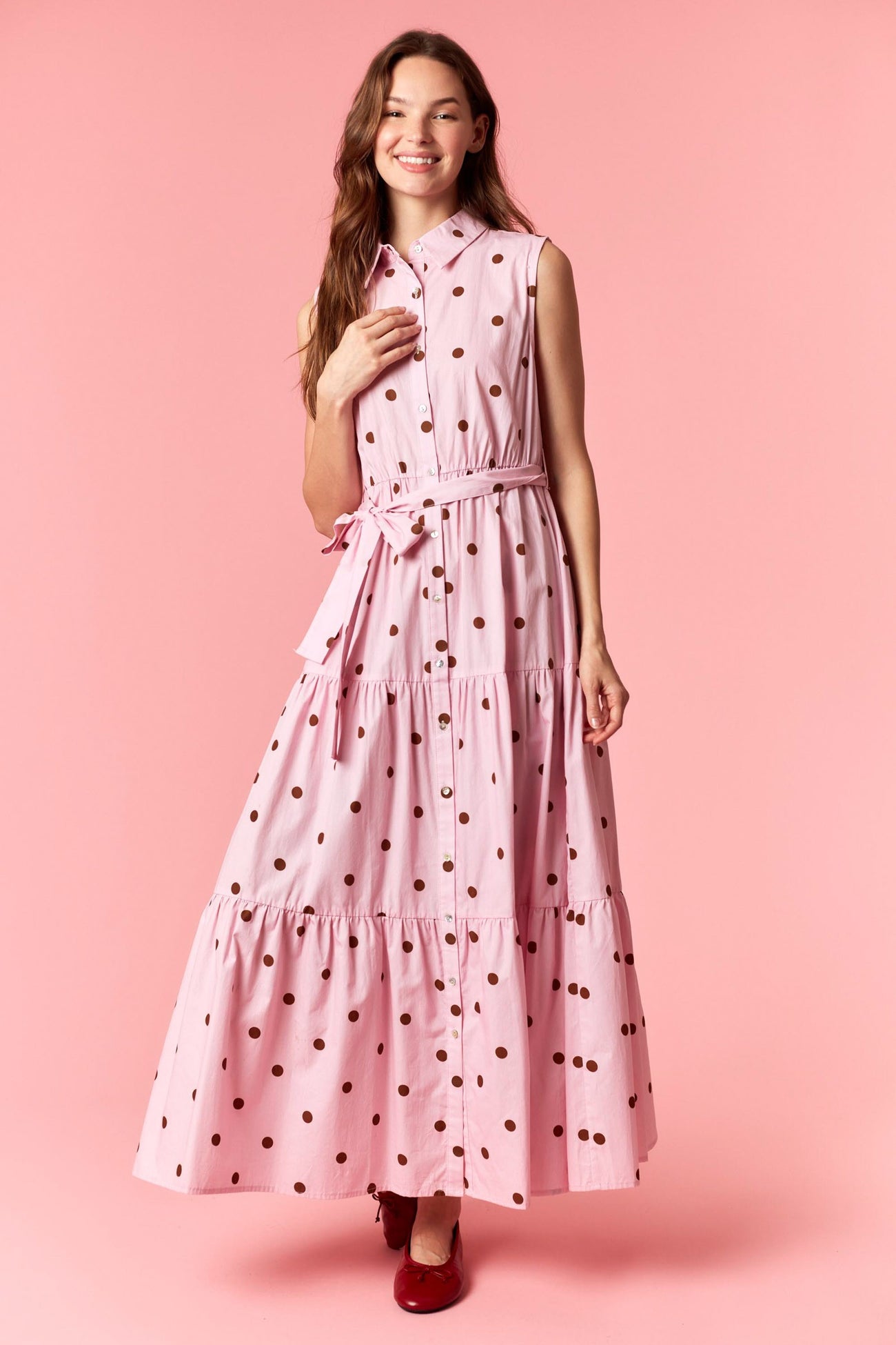 woman wearing a pink sleeveless maxi dress with brown polka dots and a tie waist detail against a pink background