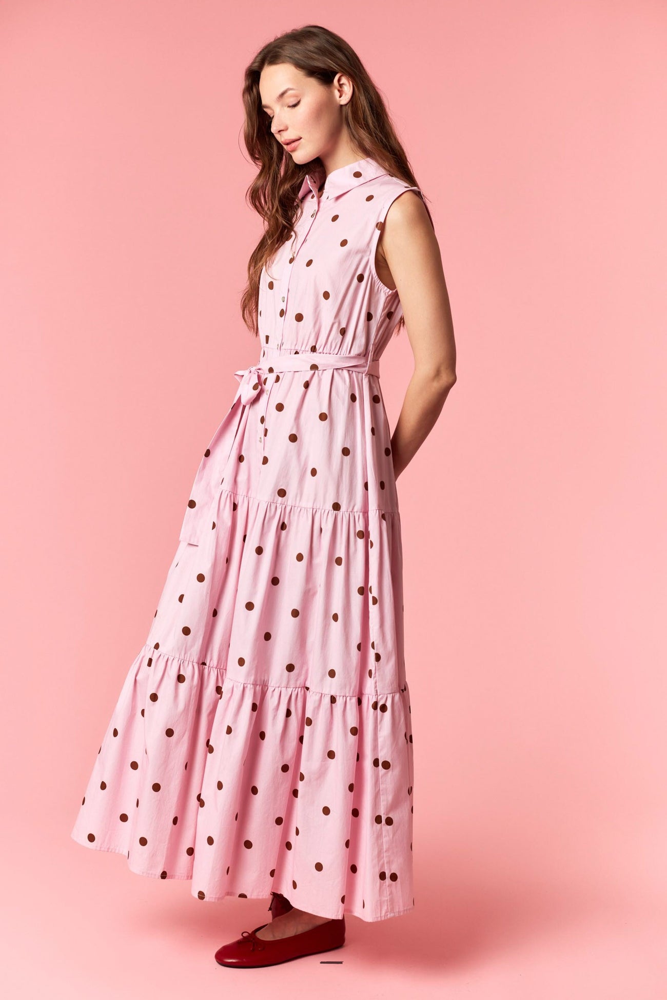 woman wearing a pink sleeveless maxi dress with brown polka dots and a waist tie, standing against a pink background