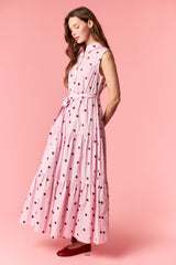 woman wearing a pink sleeveless maxi dress with brown polka dots and a waist tie, standing against a pink background